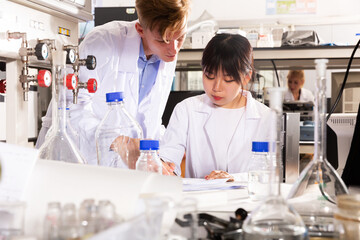 Fototapeta premium young Chinese female student with male groupmate performing experiments in university chemistry laboratory, recording results in workbook