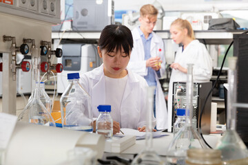 Young positive female Chinese chemist working in laboratory, writing report on results of chemical experiments in notebook