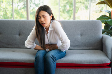 Obraz premium Unhappy Asian woman sitting on the sofa and holding on stomach suffering. Abdominal pain that comes from menstruation, diarrhea, or indigestion. Sickness and healthcare concept