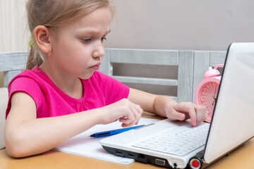 A cute little girl is sitting at a table and typing homework on a laptop to send to the teacher for review. Learning concept, distance learning, self education, video conferencing lesson with mentor