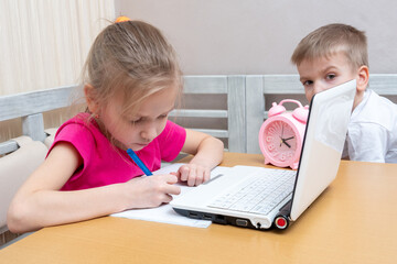 Little boy and girl study online at home. Cute little girl student writes homework in a notebook, a boy spies on the girl. Learning concept, distance learning, video conferencing lesson with mentor