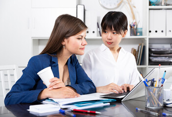 Two positive women working with laptop and papers at common table in office of international company
