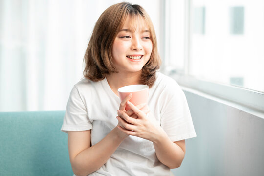 Young girl holding cup sitting by glass door in early morning