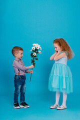 Little boy is giving to amazed little redhaired curly girl a white chrysanthemums on the blue background. Concept of happy childhood. Copy space.

