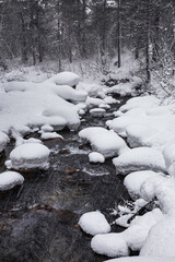 A narrow mountain river flows through a winter valley through a snow-covered forest. High caps of snow lie on stones. Heavy snowfall. Traces of flying snowflakes are visible against a dark background