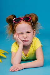 Frowning red-haired curly little girl in sunglasses is lying on the blue background with yellow plastic sun.Summer vacation and holidays concept,travel with children.Children's emotions.
