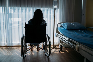 Rear view of sad and depressed female patient sitting on wheelchair with saline bottle in hospital room looking towards curtains on window