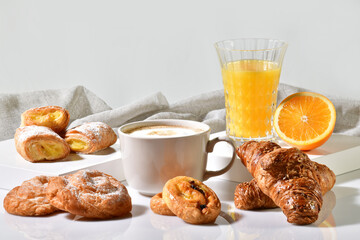 Cup of coffee surrounded by delicious pastries, with an orange juice and half orange at the back on a light background. Traditional breakfast concept.