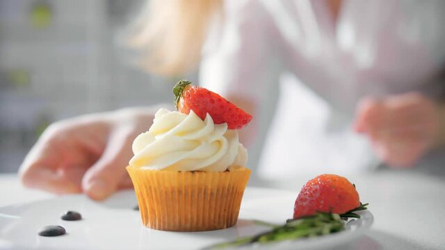 Dessert cook female hand decorates tasty cupcakes white cream topping with cutted strawberry pieces at kitchen