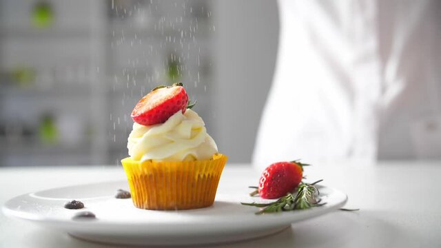 Dessert cook female hand decorates tasty cupcakes white cream topping with cutted strawberry pieces at kitchen