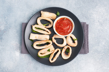 Fried squid rings with tomato sauce and lemon. Black plate, grey concrete background copy space. Top view