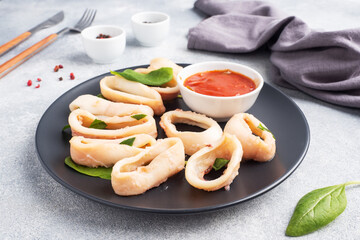 Fried squid rings with tomato sauce and lemon. Black plate, grey concrete background copy space.