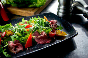 Warm salad with roast beef and fresh vegetables, served in a black plate on a marble background. Restaurant food. Close up, selective focus