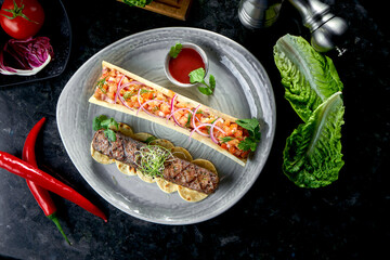 Appetizing Turkish lamb lula kebab with tomato tartare and red sauce, served in a gray plate. Dark marble background. Barbecue meat, restaurant food.