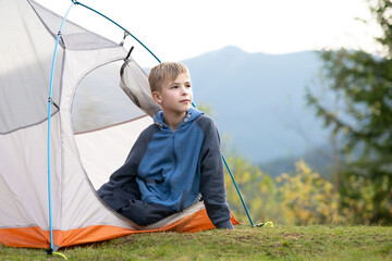 Happy hiker child boy resting in a tourist tent at mountain campsite enjoying view of beautiful summer nature. © bilanol
