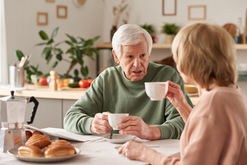 Happy senior couple sitting at the table drinking tea and talking to each other in the kitchen at home