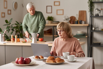 Senior woman sitting at the table with laptop and working online with her husband cooking in the background in the kitchen