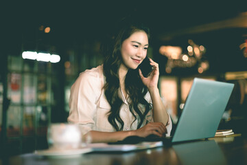 Beautiful Attractive Asian woman working with laptop smile and talking with customer in smartphone to get idea and requirement for success job outside office in coffee shop,Business Concept,Warm Tone
