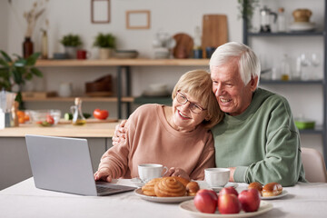 Fototapeta na wymiar Happy senior couple embracing while sitting at the table and watching movie on laptop online in the kitchen