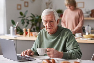 Portrait of senior man looking at camera while sitting at the table drinking tea and working on laptop with his wife in the background