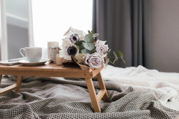 Coffee table on bed. Flowers, coffee cup and candles. Interior gray tones, plaid