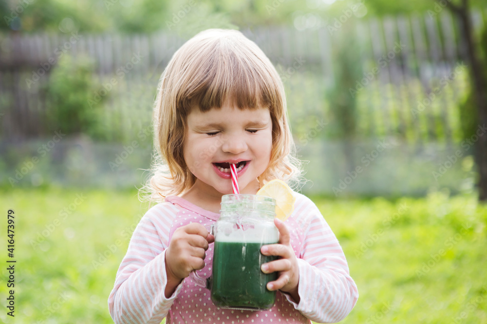 Wall mural Cute little child drinks healthy green smoothie with straw in a jar mug against the background of greenery outdoor. 