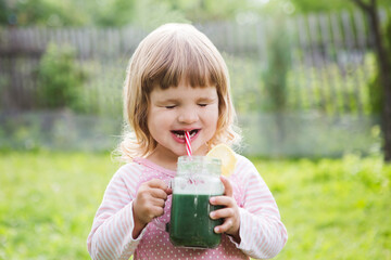 Cute little child drinks healthy green smoothie with straw in a jar mug against the background of greenery outdoor. 