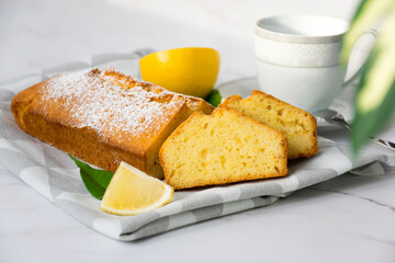 Marble table with homemade freshly baked orange loaf cake on kitchen towel, citrus fruits, cup and house plant. Traditional treat for tea or coffee. Delicious breakfast to start a day.