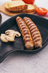 Hot juicy sausages in a frying pan with mushrooms, bread and sauce. On a stone background, top view.