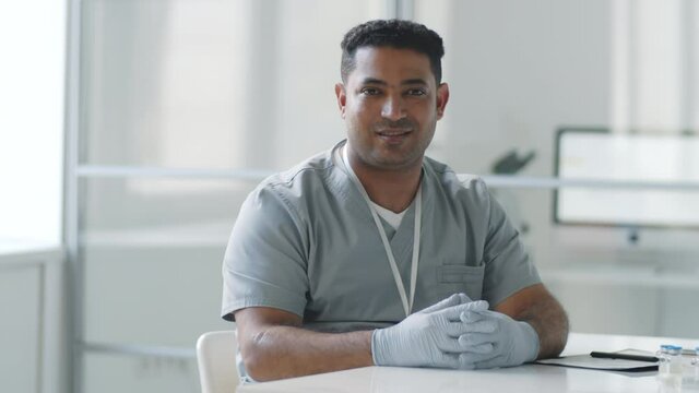Tracking shot of cheerful middle eastern male doctor in medical uniform and sterile gloves sitting at desk in clinic, looking at camera and smiling