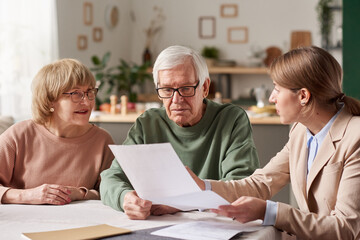 Senior couple sitting at the table and discussing documents with real estate agent they making a...