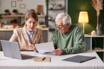 Young businesswoman consulting senior man about financial documents using laptop while they sitting at the table at home