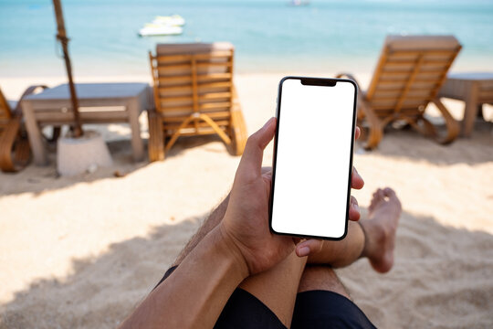 Mockup image of a man holding mobile phone with blank desktop screen while sitting on the beach