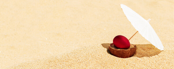 Red Easter egg on coconut shell on golden sandy beach under sun umbrella. Celebrating Easter holiday on tropical countries beach. Panoramic Easter background, wide banner, copy space