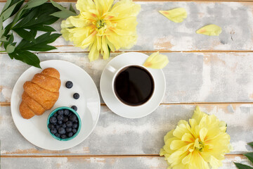 Morning coffee, croissant, blueberries and beautiful yellow peony flowers on light table top view in flat lay style. Cozy breakfast on Mother or Woman day.
