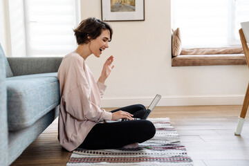 Excited beautiful girl waving hand while using laptop on floor