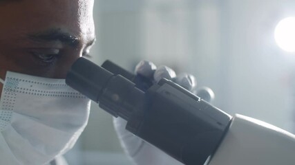 Close up tracking shot of middle eastern male scientist in protective face mask and sterile gloves looking through microscope while doing research in time of covid-19 - Powered by Adobe