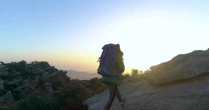 mujer escaladora con mochila y abrigo ascendiendo una monta&ntilde;a rocosa al amanecer