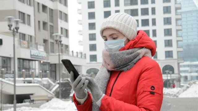 A Hipster Girl In A Red Jacket And A Protective Mask On Her Face Stands In Her Neighborhood And Writes A Message On A Black Smartphone