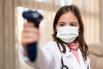 Little girl with doctor's uniform and medical mask using an infrared thermometer. Covid-19 virus safety concept.