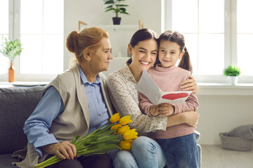 Smiling young woman thanking her little daughter and senior mom for gifts and flowers on Mother's Day. Happy beautiful multi-generational ladies celebrating Women's Day at home together