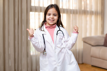 Little child dressed in doctor's uniform giving thumbs up in a fun and positive attitude. She is at home. Space for text.
