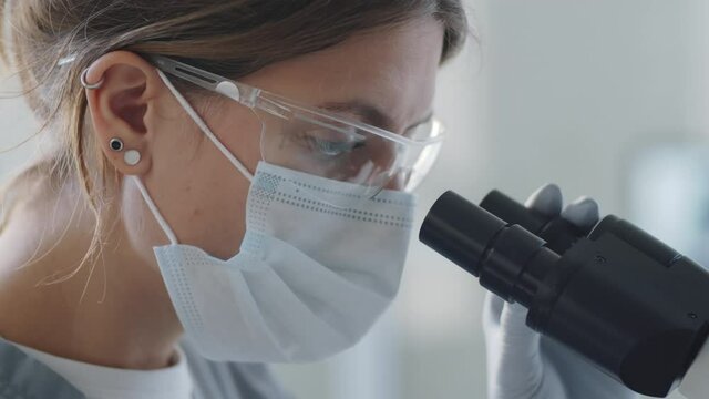 Close up shot of female lab worker in protective face mask, glasses and sterile gloves looking through microscope while conducting research in time of coronavirus pandemic