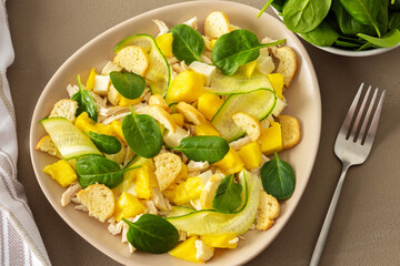 Plate with salad and fork close-up on a brown background, salad of cucumber and chicken breast, mozzarella and croutons with sweet mango pieces and spinach