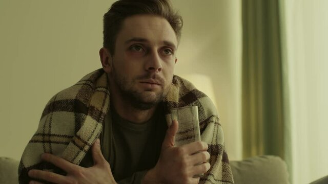 Sick man drinking glass of water with medicines, coughing and feeling fever, sitting at home wrapped in plaid