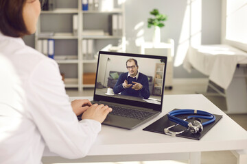 General practitioner talking to senior patient via online video conference chat. Over shoulder view laptop computer screen on desk at doctor's office. Telemedicine, virtual visit to hospital concept