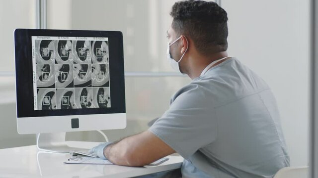Male doctor in protective face mask and gloves examining CT scan of lungs on computer and taking notes while working at desk in medical office during coronavirus pandemic