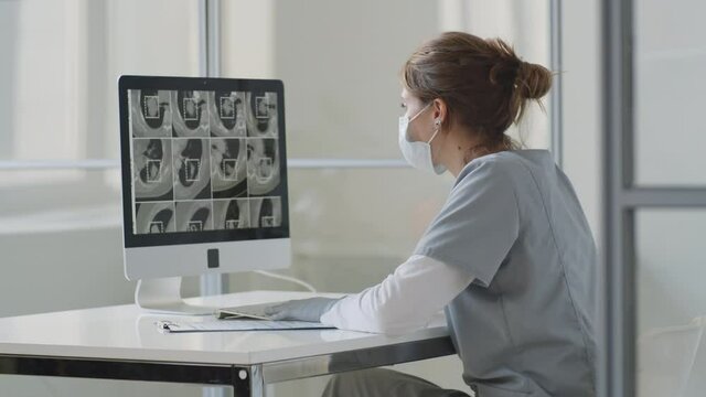 Female doctor in medical uniform, face mask and gloves examining CT scan of lungs on computer and writing on clipboard while working at desk in clinic during covid-19 outbreak