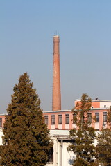 Narrow old red bricks tall industrial chimney with metal stairs mounted on side rising high above elongated factory building and trees on warm sunny winter day