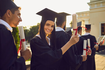 Smiling pretty girl student university graduate holding diploma with honors in hand with classmates...